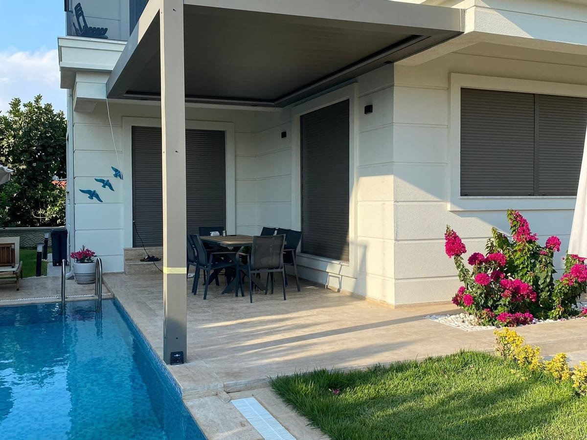 Überdachte Terrasse mit Esstisch an einem hellen Wohnhaus, davor ein Pool